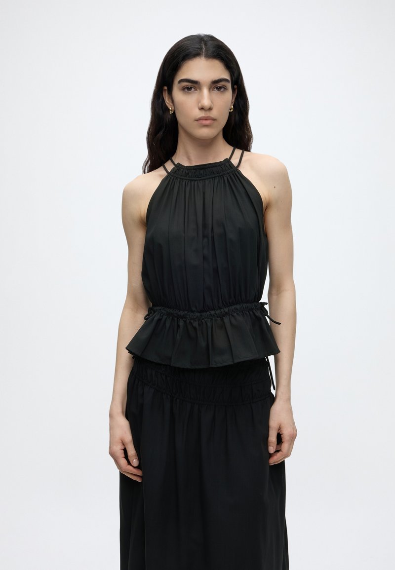 Γυναίκα με μακριά σκούρα μαλλιά που φορά μια μαύρη αμάνικη μπλούζα halter με σούρα στη μέση και μαύρη φούστα που ταιριάζει, στέκεται μπροστά σε απλό φόντο.