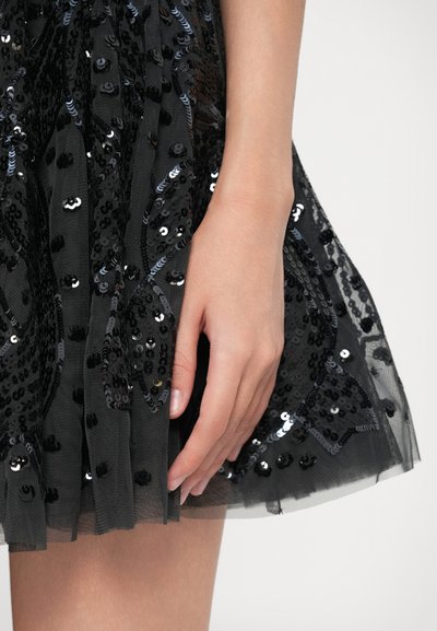 Ruka odpočívající vedle černé síťované sukně zdobené lesklými černými flitry v květinových vzorech na světlém pozadí.