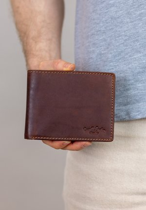 Hand holding a brown leather wallet with stitching, wearing a gray shirt and light beige pants against a neutral background.