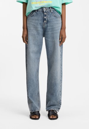 Personne portant un jean bleu ample taille haute avec braguette à boutons apparents, sandales noires à bouts ouverts et un t-shirt graphique vert clair.