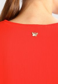 Femme portant un haut rouge texturé avec un petit broche en forme de papillon doré au dos, près de l'encolure.