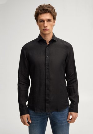 Młody mężczyzna z kręconymi włosami, ubrany w dopasowaną czarną koszulę na guziki i niebieskie dżinsy, stojący na jednolitym jasnym tle.