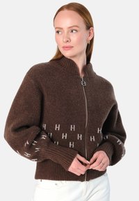 Brun zip-up cardigan med ribbet krage og mansjetter. Har et mønsterdesign med hvite 'H' bokstaver på den nedre delen. Myk tekstur.