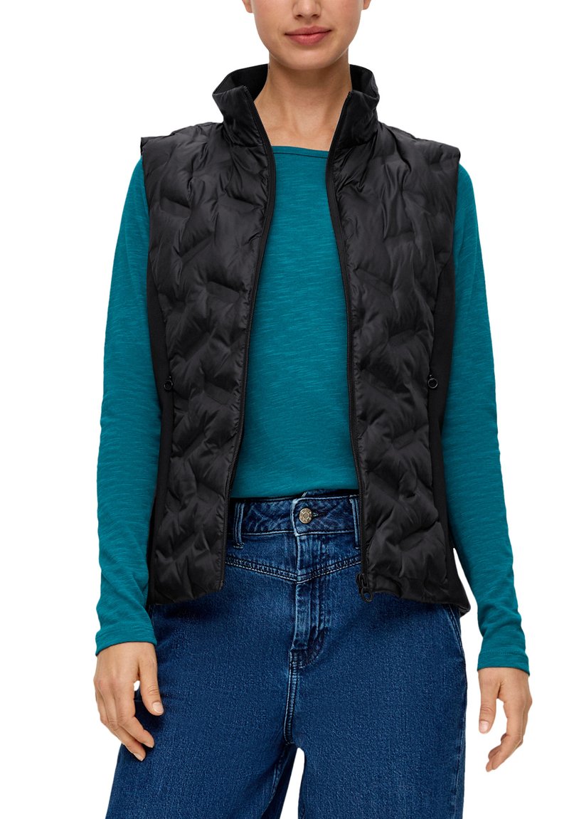 Schwarze gesteppte Weste mit hohem Kragen, mit Reißverschluss, getragen über einem türkisfarbenen Langarmshirt. Eine blaue Jeans rundet das Outfit ab.