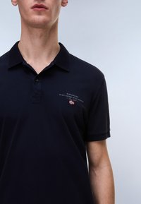 Marineblå poloshirt med krave og korte ærmer. Har broderet logo og tekst på brystet. Blødt bomuldsstof, glat tekstur.