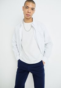T-shirt blanc sous une chemise à rayures bleues claires et blanches avec une poche poitrine, associée à un pantalon bleu marine et un collier en chaîne argenté.