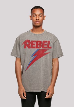 Graues, übergroßes T-Shirt mit boldem roten "REBEL"-Schriftzug und einem blauen Blitzgrafik. Der Stoff hat eine leicht abgenutzte Textur.