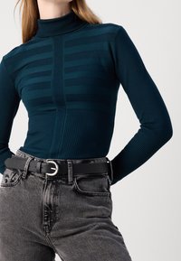 Pull à col roulé côtelé de couleur bleu-vert avec un motif rayé horizontal, associé à un jean gris foncé taille haute et une ceinture noire avec une boucle métallique.