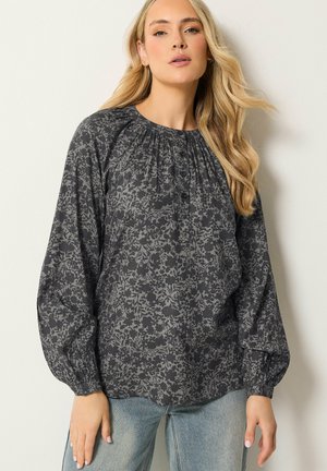 Vrouw met lang blond haar draagt een losse donkergrijze bloemenblouse met geknoopte halslijn en lichtblauwe spijkerbroek, staand tegen een witte muur.