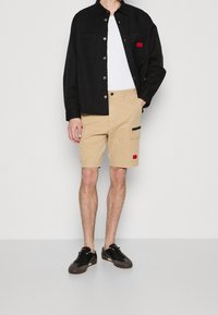 Camisa negra de botones sobre una camiseta sin mangas blanca, combinada con pantalones cortos beige que tienen un bolsillo con cremallera y una etiqueta roja, usados con zapatillas negras.