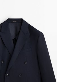 Blazer bleu marine croisé avec revers pointus et quatre boutons noirs sur fond blanc.