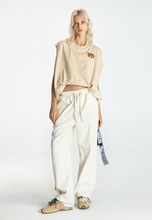 Mujer rubia con top beige sin mangas con parche de oso de peluche, pantalones blancos sueltos con cordón y zapatillas beige con cordones de diferentes colores.