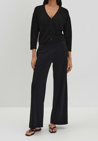 Černý žebrovaný kardigan s výstřihem do V a zapínáním na knoflíky, spárovaný s volnými černými kalhotami, obě vyrobené z hladké látky.