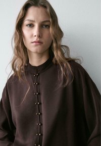 Braunes Oberteil mit hohem Kragen und Knopfleiste mit runden Verzierungen, gefertigt aus einem weichen, strukturierten Stoff.