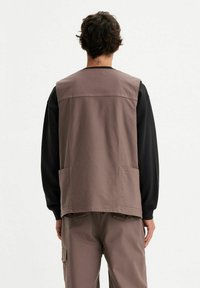 Gilet marrone in tela con taglio dritto, due tasche frontali e yoke posteriore. Indossato sopra una camicia nera a maniche lunghe con una texture morbida.