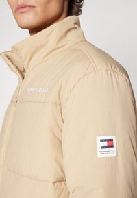 Beige puffer jacket with a high collar, featuring horizontal quilted texture, and a logo patch on the left sleeve.