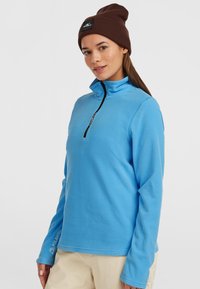 Lyseblå fleece pullover med halvhøj krave og lynlås, lange ærmer og en subtil logo detalje. Blød tekstur. Båret med en brun hue.