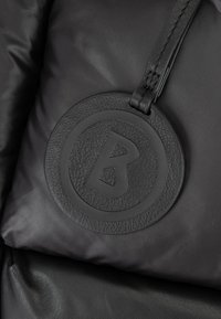 Črna puhasta jakna z teksturirano usnjeno oznako, ki vključuje krožni emblem in izstopajoč logotip "B", pritrjena z našitjem.