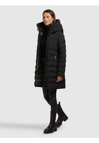 Kvinde iført en lang, sort quiltet frakke med høj krave, sort rullekrave, læderbukser og sorte chunky støvler, stående på en ensartet baggrund.