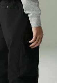 Schwarze Cargo-Hosen mit Seitentaschen, die über eine glatte Textur verfügen. Eine Nahaufnahme einer Hand, die auf der Tasche ruht, ist zu sehen. Die Bündchen eines grauen Sweatshirts sind sichtbar.