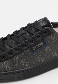 Svart sneaker med texturerad tyg som har ett monogrammönster, prydd med svarta snören och ett logomärke i blå och silverfärgade detaljer.
