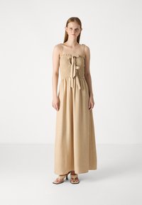Beige maxi-jurk met een gesmockte buste, ruches en verstelbare spaghettibandjes. Heeft een strikdetail aan de voorkant en losvallende, vloeiende stof.