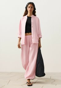 Femme portant un pantalon rose ample et une chemise par-dessus un crop top noir, tenant un grand sac noir texturé, debout sur un sol clair carrelé.