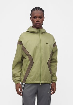 Man met gevlochten haar die een lichtgroene capuchontrui met rits draagt met bruine accenten en grijze broek met witte strepen en een logo.