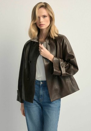 Blondynka w luźnej ciemnobrązowej skórzanej kurtce na białym topie i niebieskich dżinsach, trzymająca kołnierz kurtki jedną ręką.