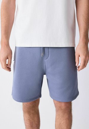 Mann, der hellblaue Shorts mit Zugband und ein weißes T-Shirt trägt, von der Taille bis zur Mitte des Oberschenkels vor einem einfarbigen Hintergrund gezeigt.