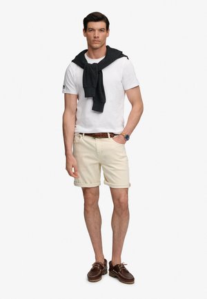 Ung mand iført hvid t-shirt, beige op-rullede shorts, brun bælte og sko, sort sweater over skuldrene og ur på venstre håndled.