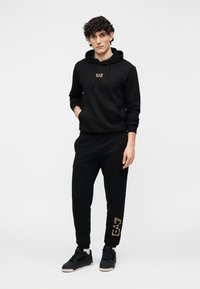 Juodas medvilninis džemperio su gobtuvu ir treniruočių kostiumo komplektas. Džemperis turi mažą logotipą ir kišenę kiaurymę; kelnėse plėtyje yra auksinio spausdinimo detalė.