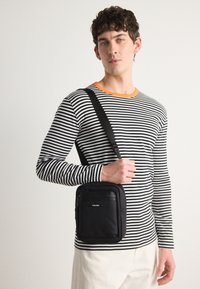 Borsa a tracolla nera con texture liscia, dotata di una tasca frontale con cerniera e dettaglio del logo. Abbinata a una camicia a maniche lunghe a righe.