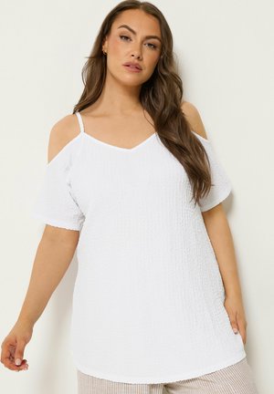Kvinde med langt brunt hår iført en tekstureret hvid cold-shoulder top og stribede beige bukser mod en lys baggrund.