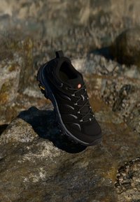 Black hiking shoe with a mesh upper and rubber sole, highlighted by gray accents and a yellow logo, placed on a rocky surface.