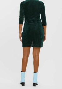 Vestido de terciopelo acanalado verde con mangas tres cuartos, que presenta un diseño ajustado y una longitud a mitad del muslo. Combinado con botas de tobillo azul claro.