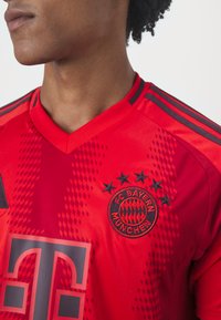 Rød FC Bayern München-drakt med V-hals, med svart logo og tekst, teksturert mønster, og kontrasterende svarte detaljer på skuldrene.
