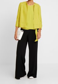 Vihreä limen värinen lyhyt takki, yhteensopiva yläosa, mustat leveälahkeiset housut ja valkoinen laukku. Sujuva rakenne, jämäkkä muotoilu, minimalistiset yksityiskohdat.