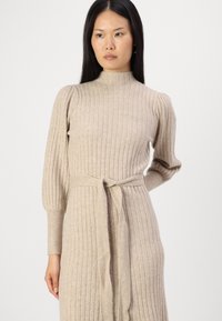 Robe en tricot beige avec une texture côtelée, col montant, manches bouffantes et ceinture à la taille. Le tissu semble doux et légèrement structuré.