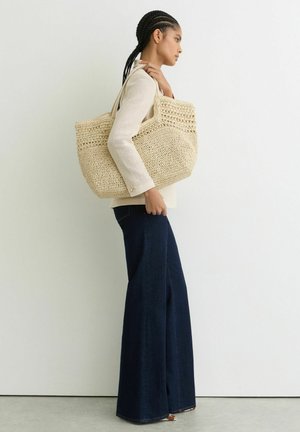 Žena s copánky, oblečená v béžovém topu a tmavých širokých džínách, s velkou béžovou pletenou kabelkou přes rameno.