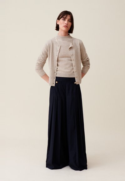 Cardigan en tricot beige sur un pull à col roulé beige, associé à des pantalons larges bleu marine avec des boutons dorés.