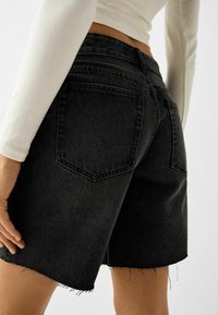 Pantalones cortos de mezclilla negra con dobladillo deshilachado, que cuentan con dos bolsillos traseros y un diseño de cintura alta. La tela tiene una textura suave.