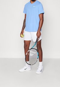Blå Nike V-ringad t-shirt, vita shorts, vita Nike-skor, håller en gul tennisboll och ett racket med blå accenter.