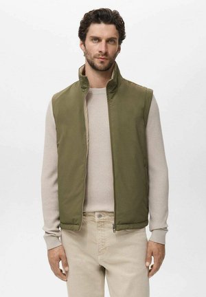 Olive green sleeveless vest with a high collar, textured interior, front zipper, paired with a light beige ribbed sweater and pants.