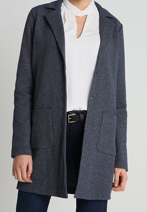 Personne portant un long manteau gris foncé sur un chemisier blanc avec une encolure en trou de serrure et un pantalon foncé avec une ceinture noire.