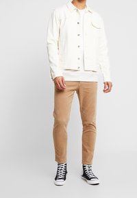 Biała kurtka dżinsowa na guziki, beżowe spodnie sztruksowe oraz czarne trampki za kostkę. Prosty design i neutralna paleta kolorów w całym zestawie.