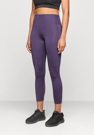 Kobieta ubrana w fioletowe legginsy z wysokim stanem i czarne buty sportowe, stoi na jednolitym jasnym tle.