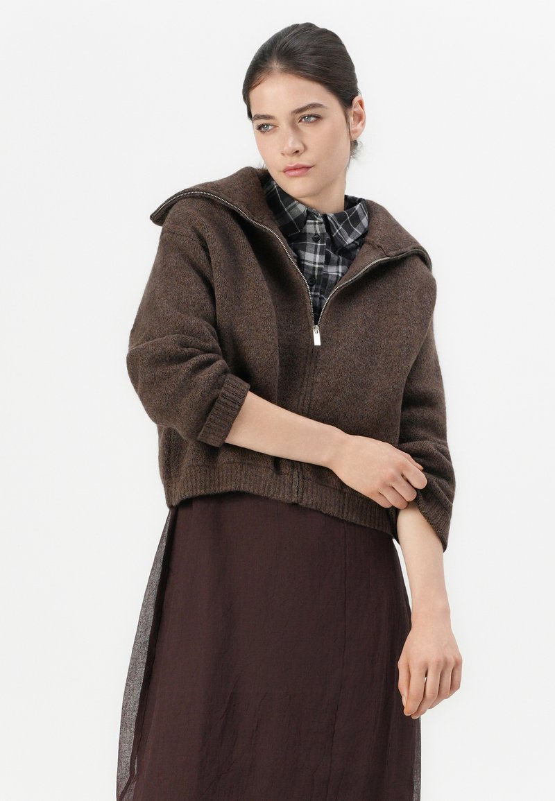 Bruine gebreide sweater met een ritskraag, cropped design en geribbelde mouwen. Gedragen over een donkere, vloeiende rok. Zachte textuur, casual pasvorm.