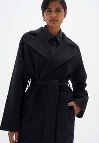 Manteau trench en laine noir avec de larges revers, manches longues et une ceinture nouée à la taille. Il dispose de poches latérales et d'une texture douce.