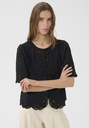 Tânără cu păr șaten deschis, purtând o bluză neagră cu mâneci scurte brodată și pantaloni de culoare crem, cu brațele încrucișate, fundal neutru.